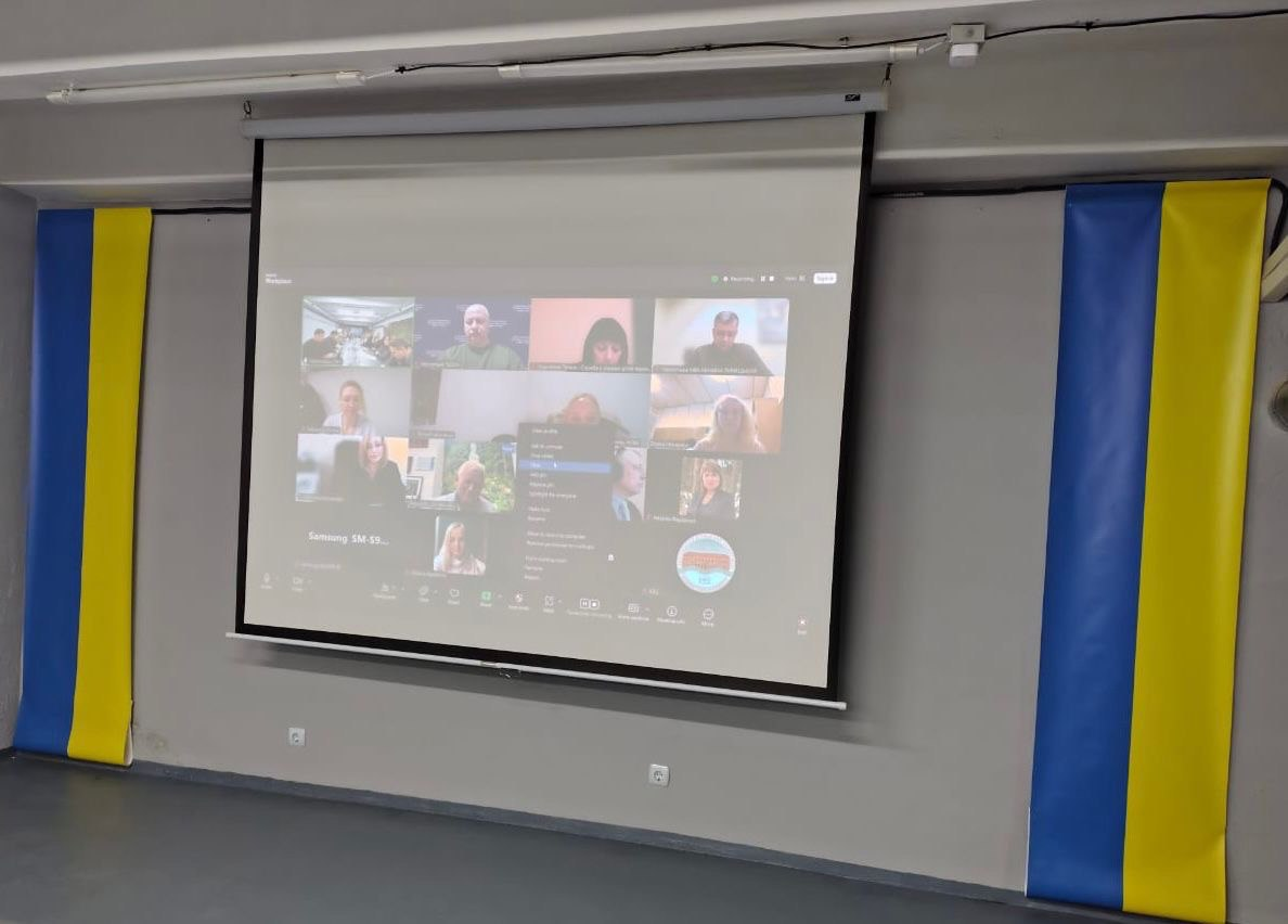 Проєкційний екран на стіні, на якому транслюється онлайн-нарада з багатьма учасниками. По боках від екрана розміщені два вертикальні прапори України.