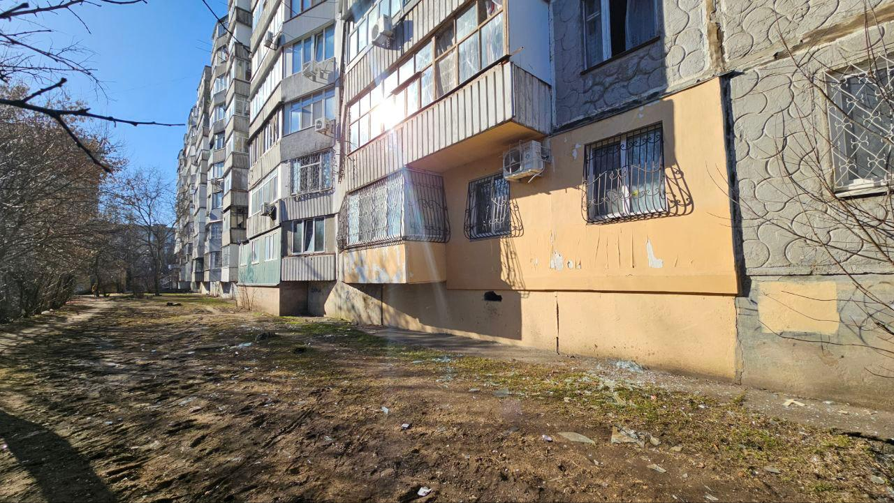 Багатоповерховий житловий будинок із заскленими балконами та решітками на вікнах першого поверху. Перед будівлею — прибудинкова територія з деревами без листя у сонячний день.