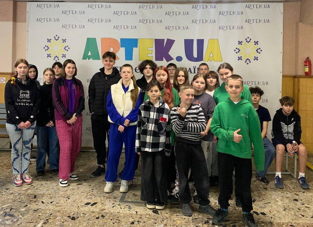 Група підлітків позує для спільного фото в приміщенні на фоні банера з написом «ARTEK.UA». Діти одягнені у повсякденний та спортивний одяг різних кольорів.