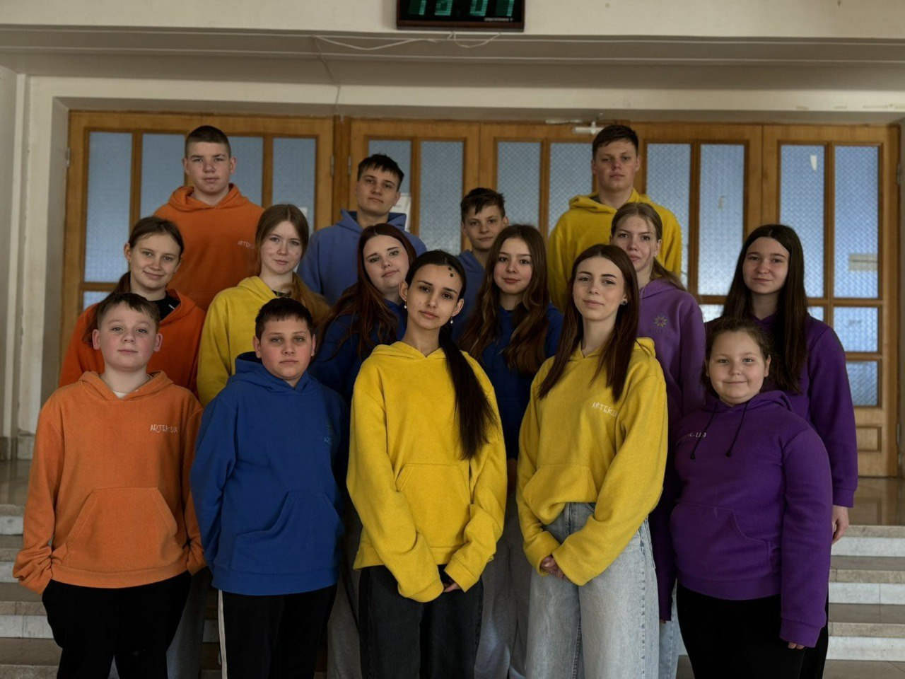 Група дітей та підлітків позує для спільного фото в приміщенні на фоні дерев'яних дверей. Учасники одягнені в однотипні худі яскравих кольорів: жовтого, синього, фіолетового та помаранчевого.