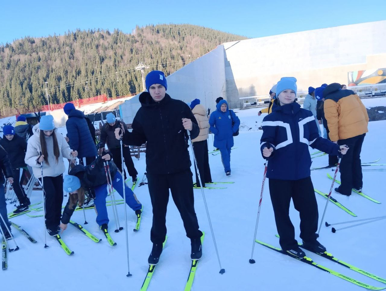 Група підлітків на лижах на засніженому тренувальному майданчику в горах. Вони одягнені в теплий спортивний одяг та шапки, на фоні видно схил із хвойним лісом та бетонні конструкції.