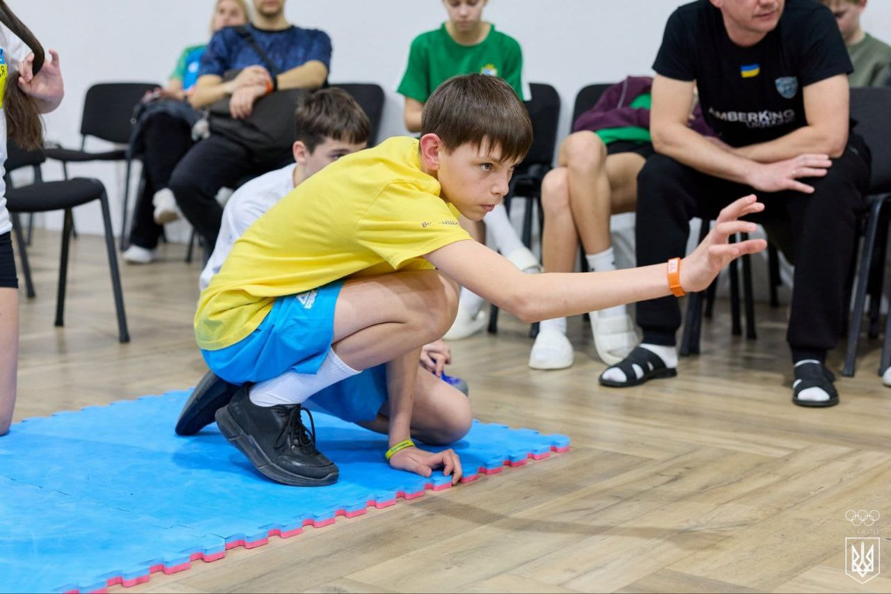 Хлопець у жовтій футболці та блакитних шортах зосереджено виконує вправу, присівши на край синього спортивного мату. На задньому плані сидять інші підлітки та дорослий, спостерігаючи за процесом у спортивному залі.