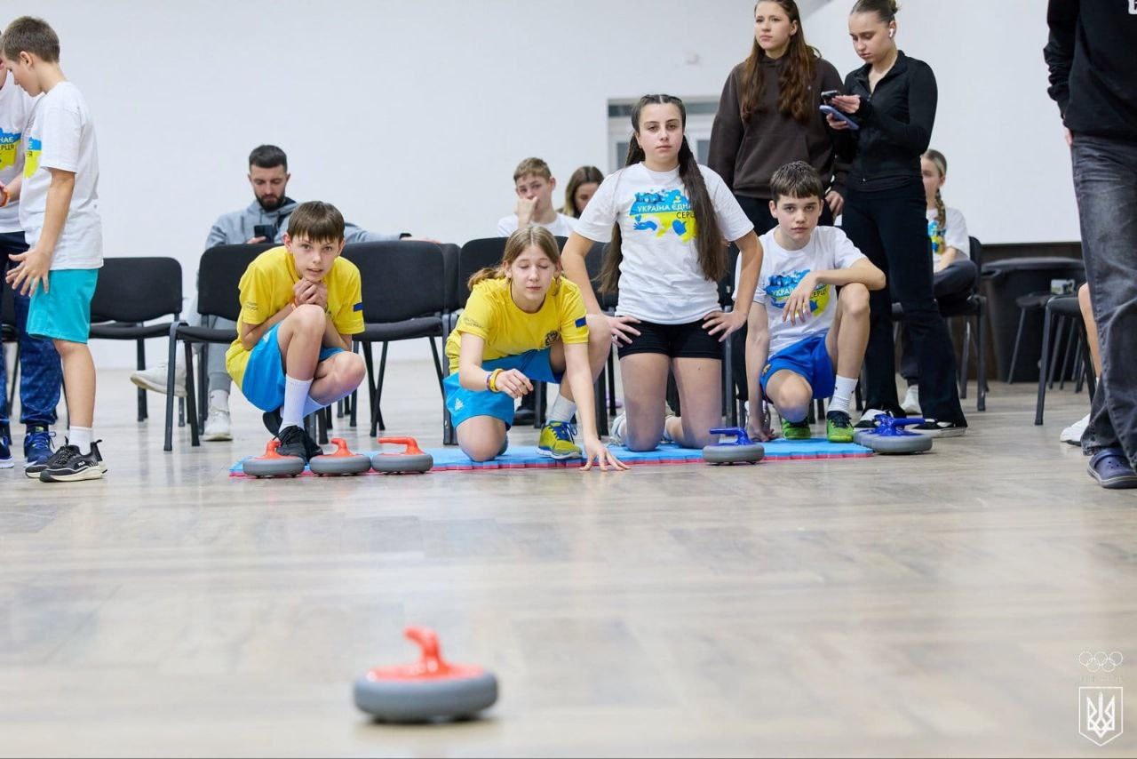 Група підлітків у жовто-блакитній спортивній формі грає в підлоговий керлінг у залі. На передньому плані по підлозі котиться камінь, а гравці зосереджено спостерігають за грою з матів.