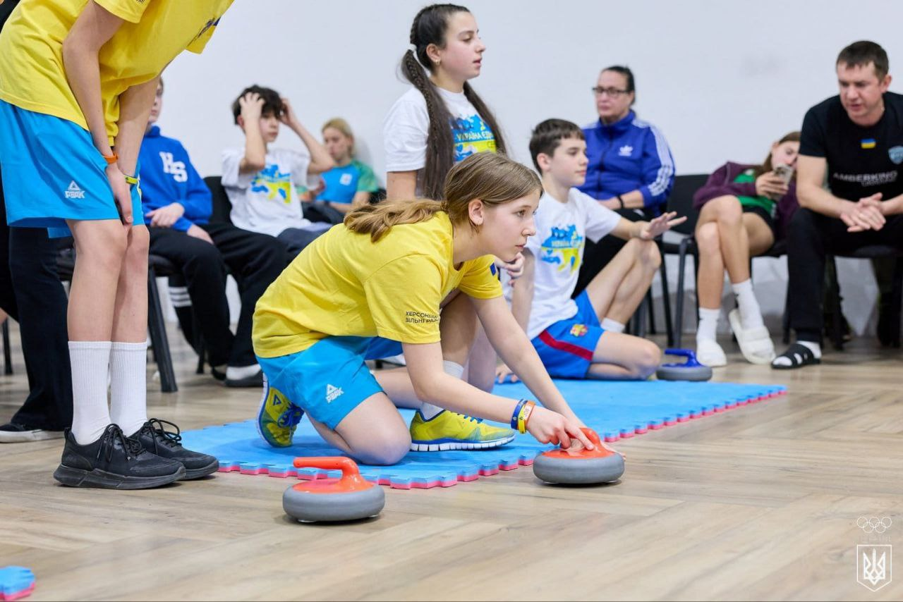 Дівчина в жовтій футболці та блакитних шортах зосереджено готується до кидка каменю для підлогового керлінгу. Вона перебуває на синьому спортивному маті, а на задньому плані за грою спостерігають інші підлітки та тренер.