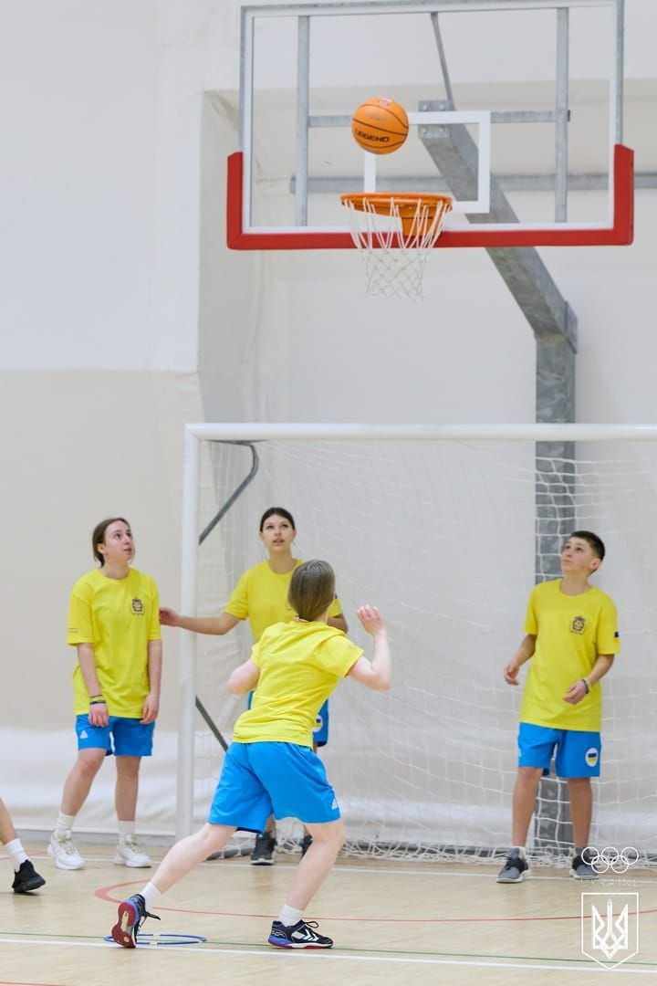 Група підлітків у жовто-блакитній спортивній формі грає в баскетбол у спортзалі. М'яч знаходиться в повітрі над кошиком, а гравці спостерігають за його польотом.