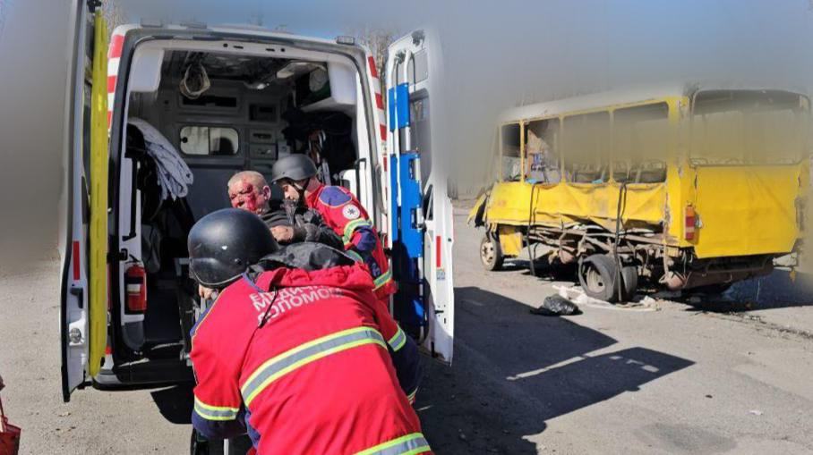 Рятувальники та медики надають допомогу пораненому чоловікові біля відчинених дверей реанімобіля. Поруч на дорозі стоїть сильно пошкоджений жовтий автобус із вибитим склом та деформованим кузовом.