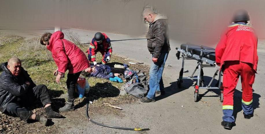 На місці події медики та очевидці надають допомогу постраждалим. На узбіччі сидить чоловік із травмами обличчя, інша людина лежить на землі під наглядом рятувальника, поруч підготовлені медичні ноші.