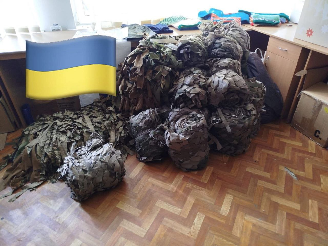 Рулони маскувальних сіток на паркетній підлозі в приміщенні. У верхньому лівому куті додано емодзі прапора України.