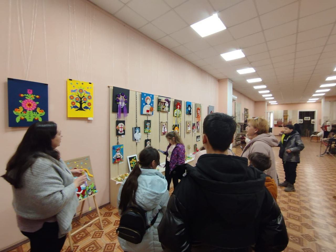 Група людей, серед яких дорослі та діти, розглядають виставку дитячих творчих робіт у світлому коридорі. На стінах та спеціальних стендах розміщено різнокольорові аплікації, малюнки та пороби, виконані в різних техніках, зокрема з використанням народних мотивів.