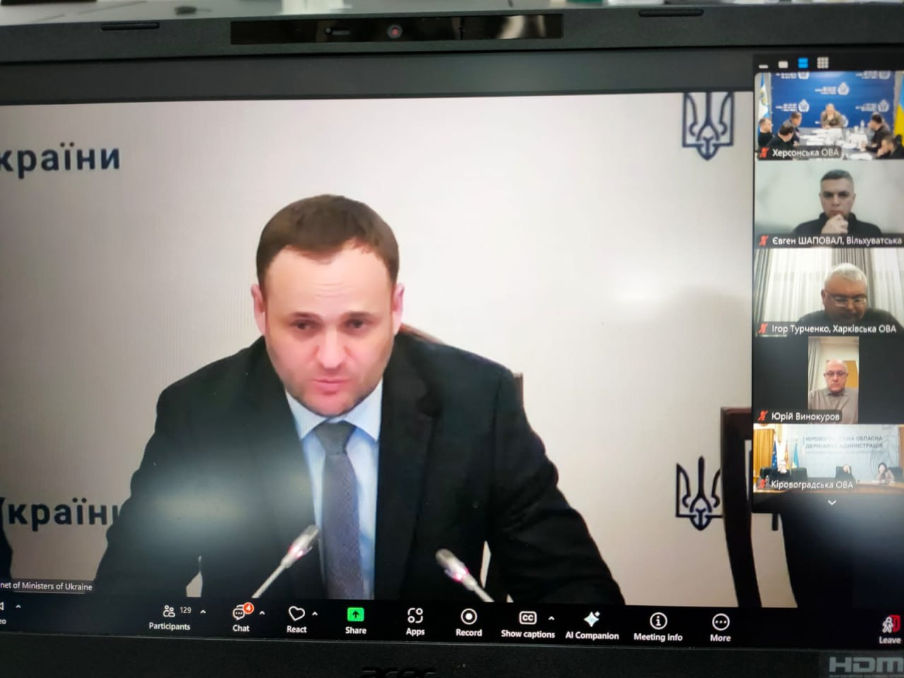 Скриншот екрана ноутбука під час онлайн-наради. На передньому плані — головний доповідач у костюмі, праворуч у колонці відображаються інші учасники відеоконференції.