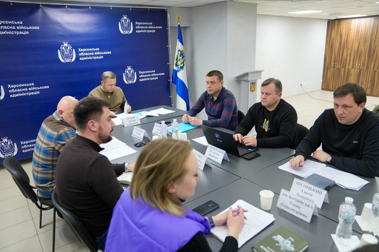 Робоча зустріч посадовців у кабінеті. Учасники сидять за спільним столом із документами та ноутбуком на фоні синіх інформаційних стендів та прапора.