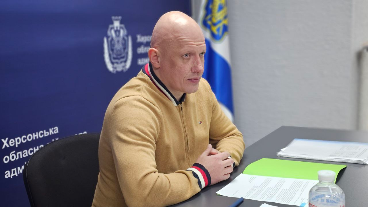 Офіційний представник за робочим столом на фоні державного прапора та банера з офіційною символікою.