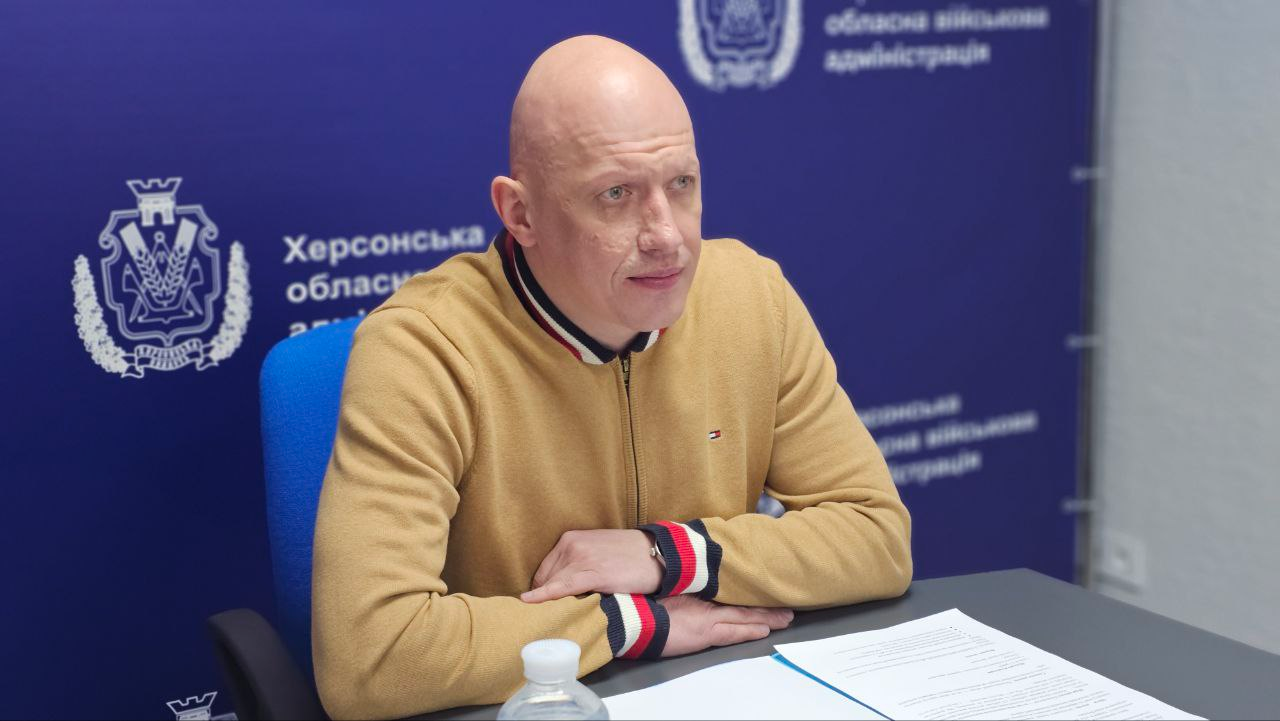 Чоловік у бежевому джемпері сидить за столом під час офіційної зустрічі. Він дивиться вбік, на столі перед ним лежать документи. Позаду — синє офіційне тло з гербами та написами.