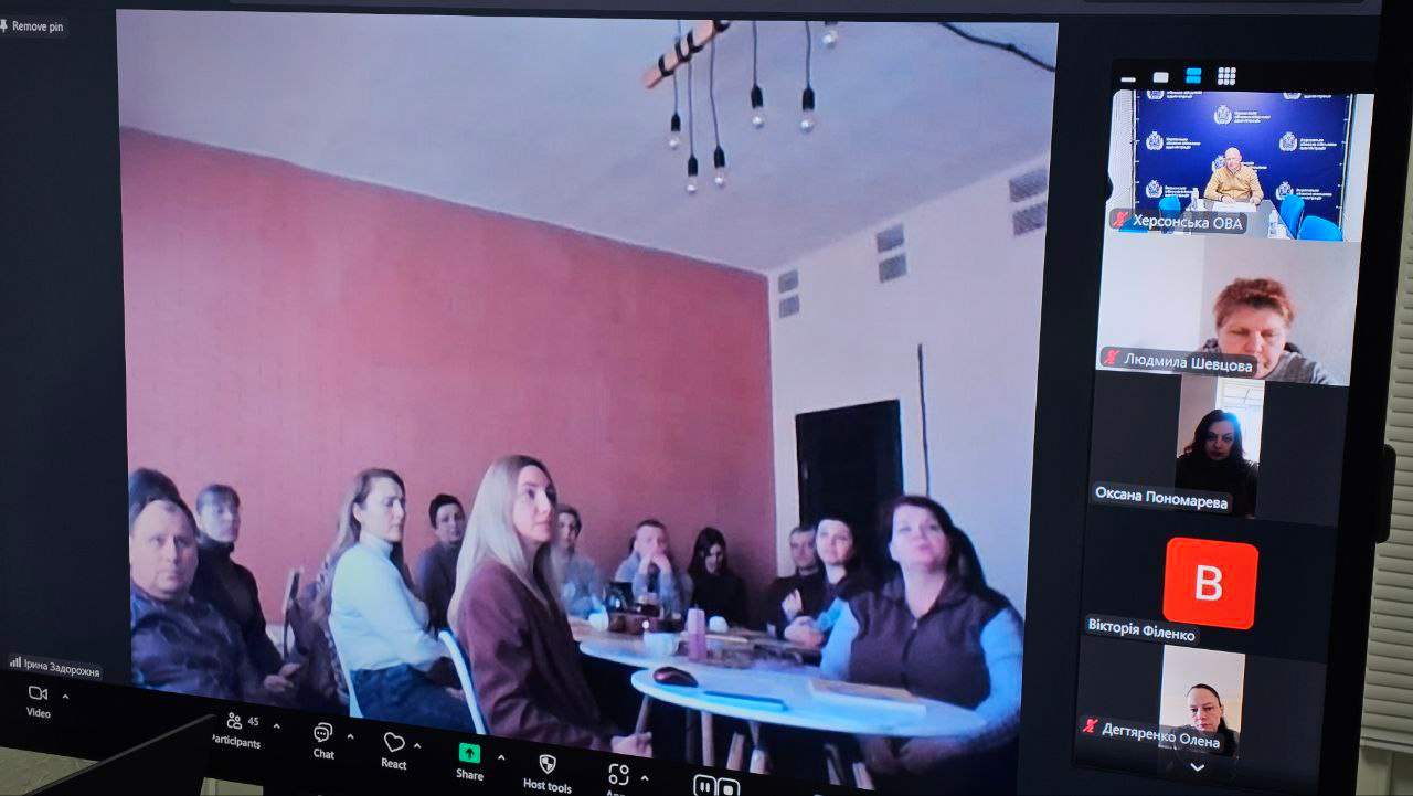 Скриншот відеоконференції у Zoom. На головному екрані — група людей в офісному приміщенні, які слухають доповідача. На бічній панелі відображено вікна інших учасників, що підключилися дистанційно.