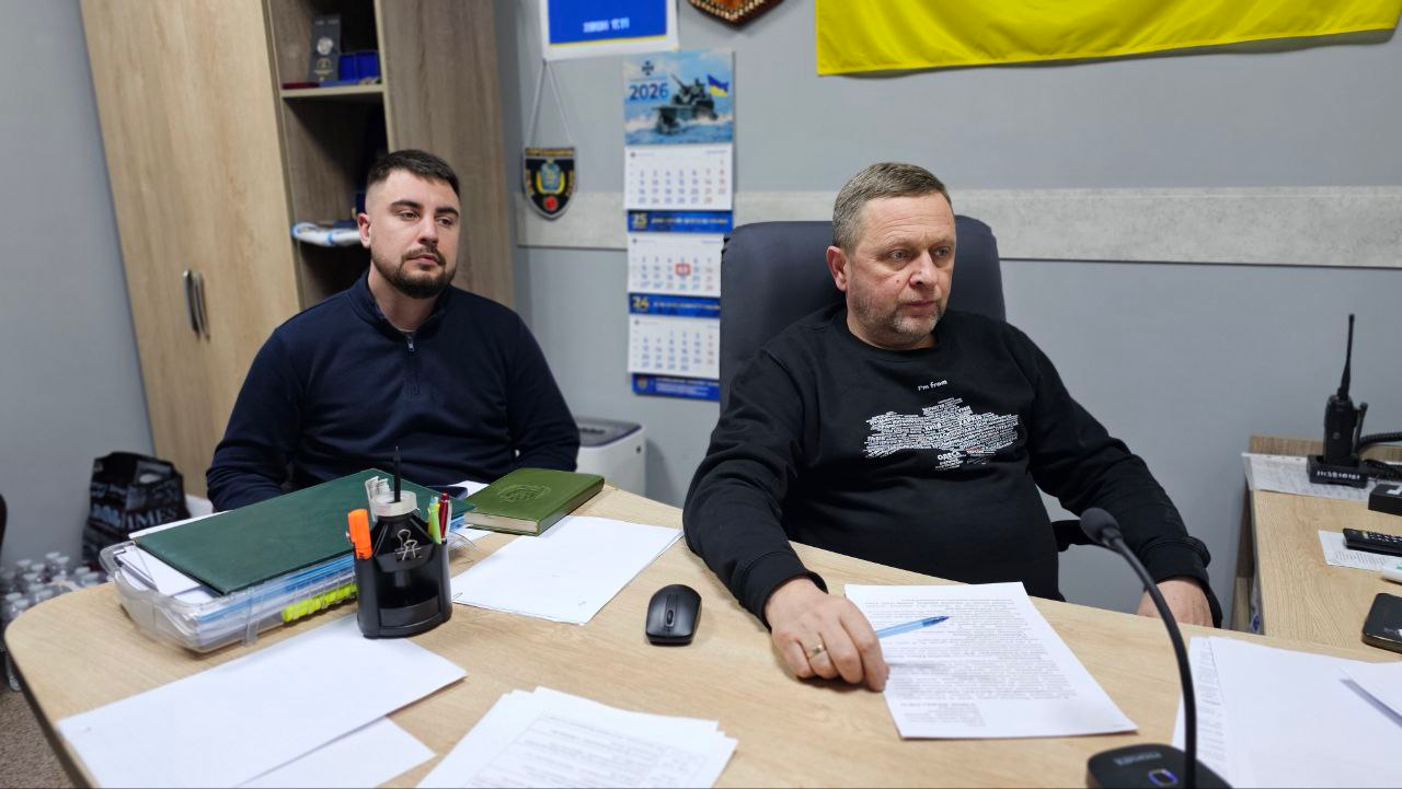 Двоє чоловіків сидять за робочим столом у кабінеті. Один із них, у чорному світшоті з принтом карти України, тримає ручку та дивиться вперед. Інший, у темно-синьому одязі, сидить поруч. На столі розкладені документи, канцелярське приладдя та комп'ютерна миша. На стіні позаду висять календар і частина прапора України.