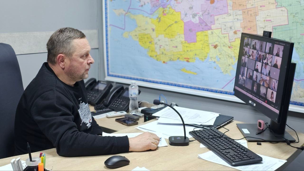 Чоловік за робочим столом проводить відеоконференцію в офісі. На стіні за ним розміщена велика географічна карта, а на столі — монітор, документи, мікрофон та стаціонарні телефони.