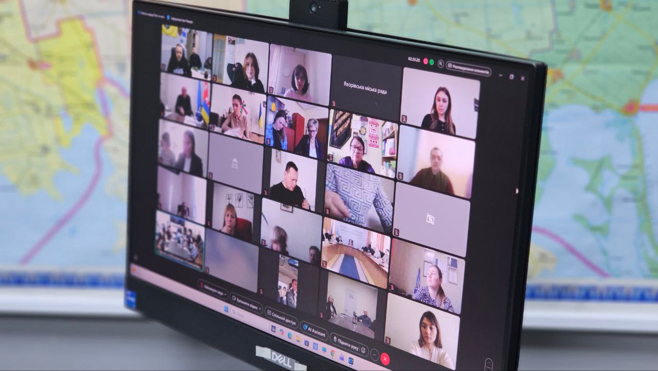 Монітор комп'ютера, на якому відображено онлайн-нараду з багатьма учасниками у форматі відеосітки. На задньому плані за монітором видно частину великої карти.