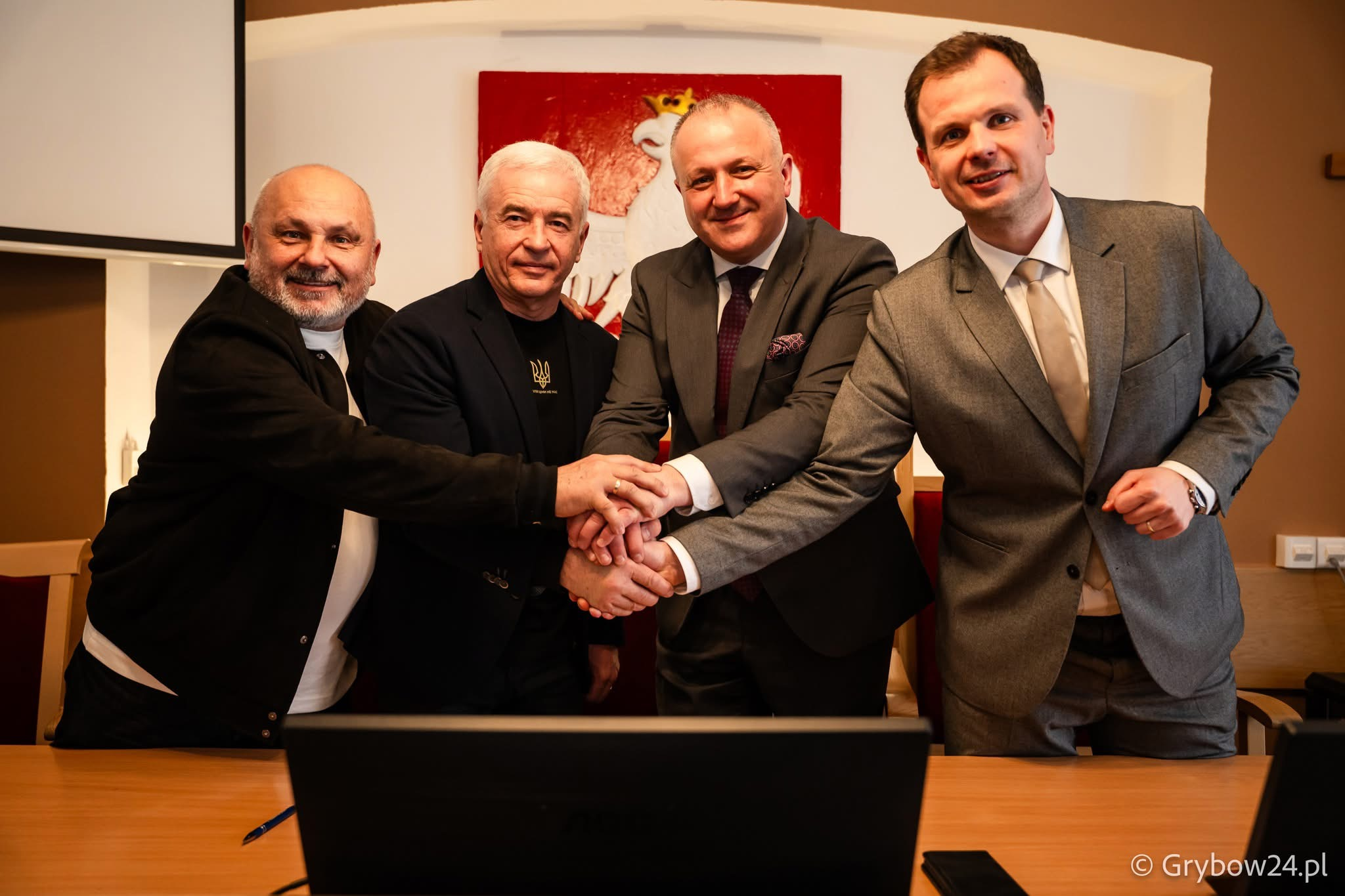 Четверо чоловіків у діловому та кежуал одязі стоять за столом і разом тиснуть один одному руки. На фоні видно герб Польщі, а на одязі одного з чоловіків — малий герб України.