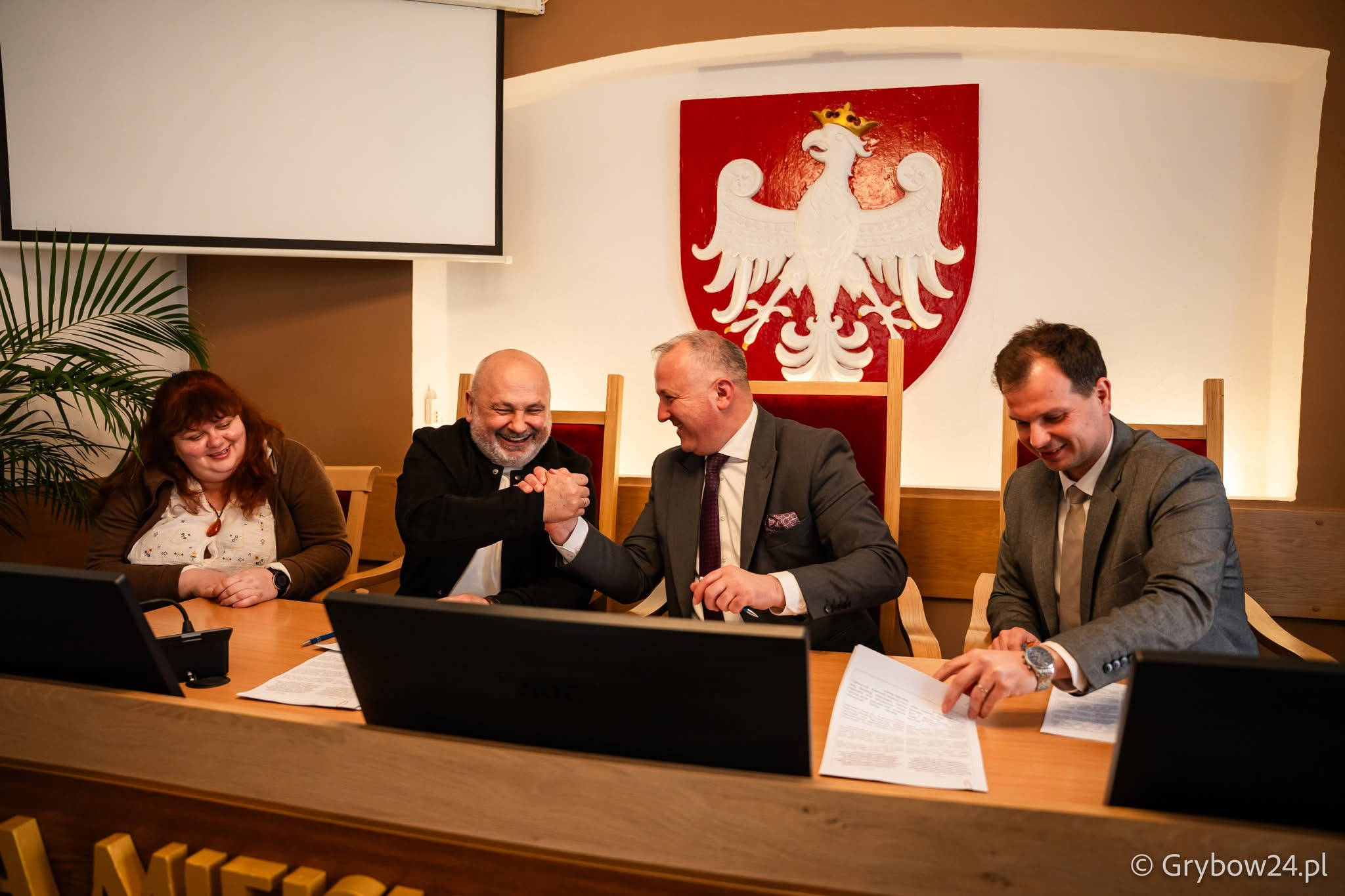 Четверо людей сидять за довгим столом в офіційному приміщенні. Двоє чоловіків у центрі посміхаються та тиснуть один одному руки, інші двоє — жінка та чоловік — дивляться на документи або спостерігають за моментом. На стіні позаду розміщено герб Польщі.