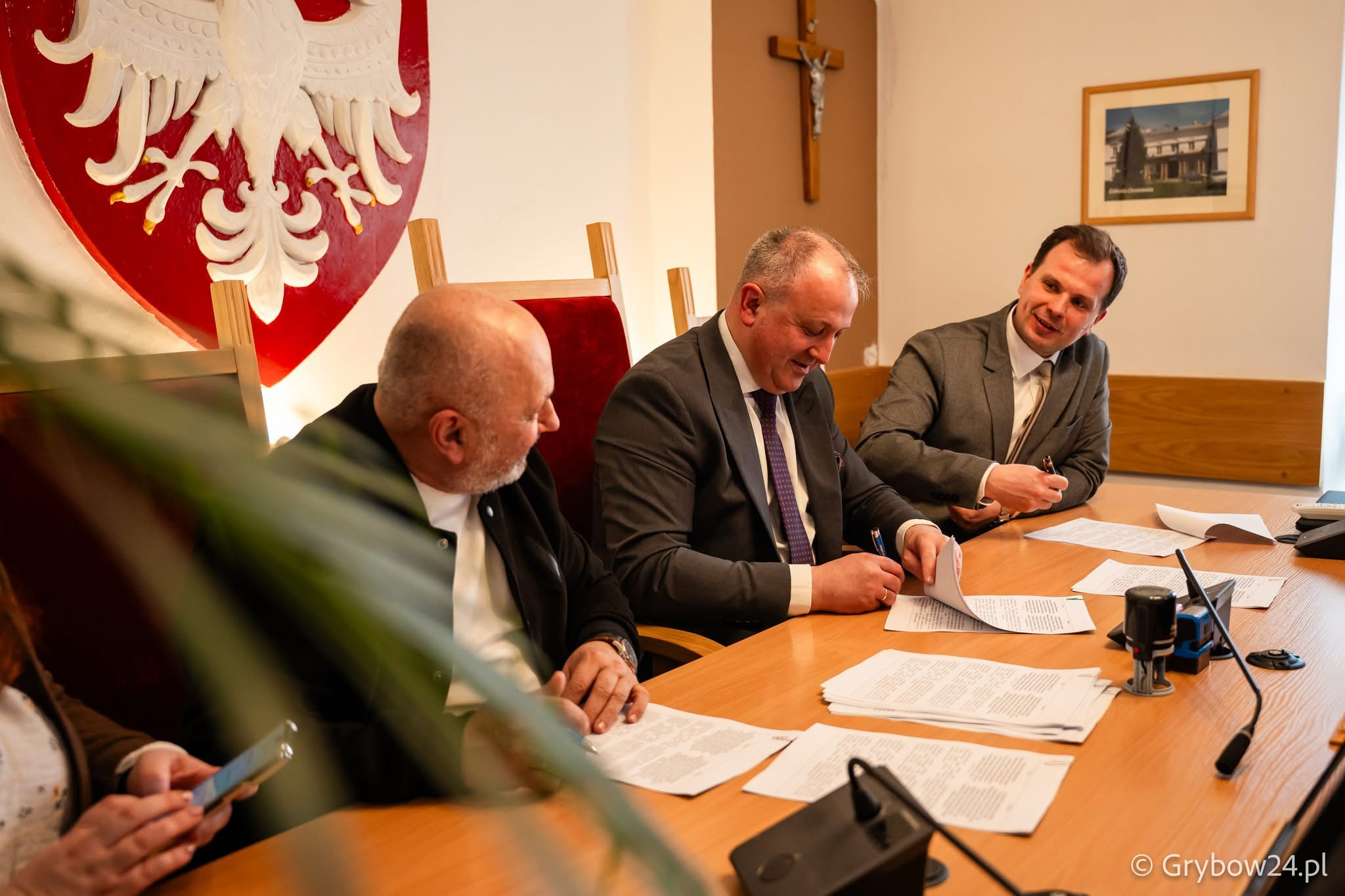 Четверо людей сидять за довгим столом під час офіційної зустрічі. Троє чоловіків у центрі підписують та обговорюють документи, а на передньому плані ліворуч видно частину постаті четвертої особи зі смартфоном. На стіні позаду розміщено великий герб Польщі та розп'яття.