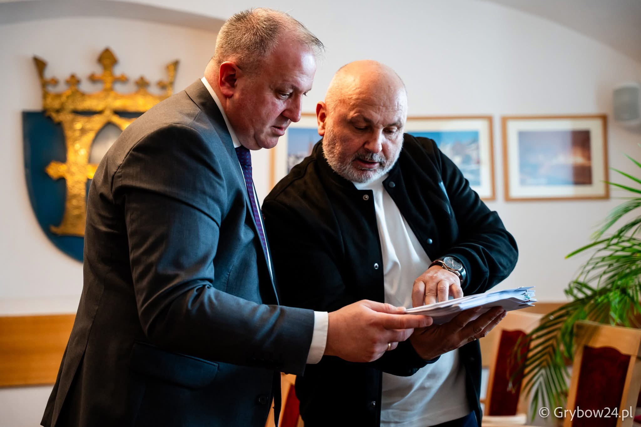 Двоє чоловіків у діловому та повсякденному одязі зосереджено розглядають документи в папці. На стіні позаду видно герб і картини в рамах.