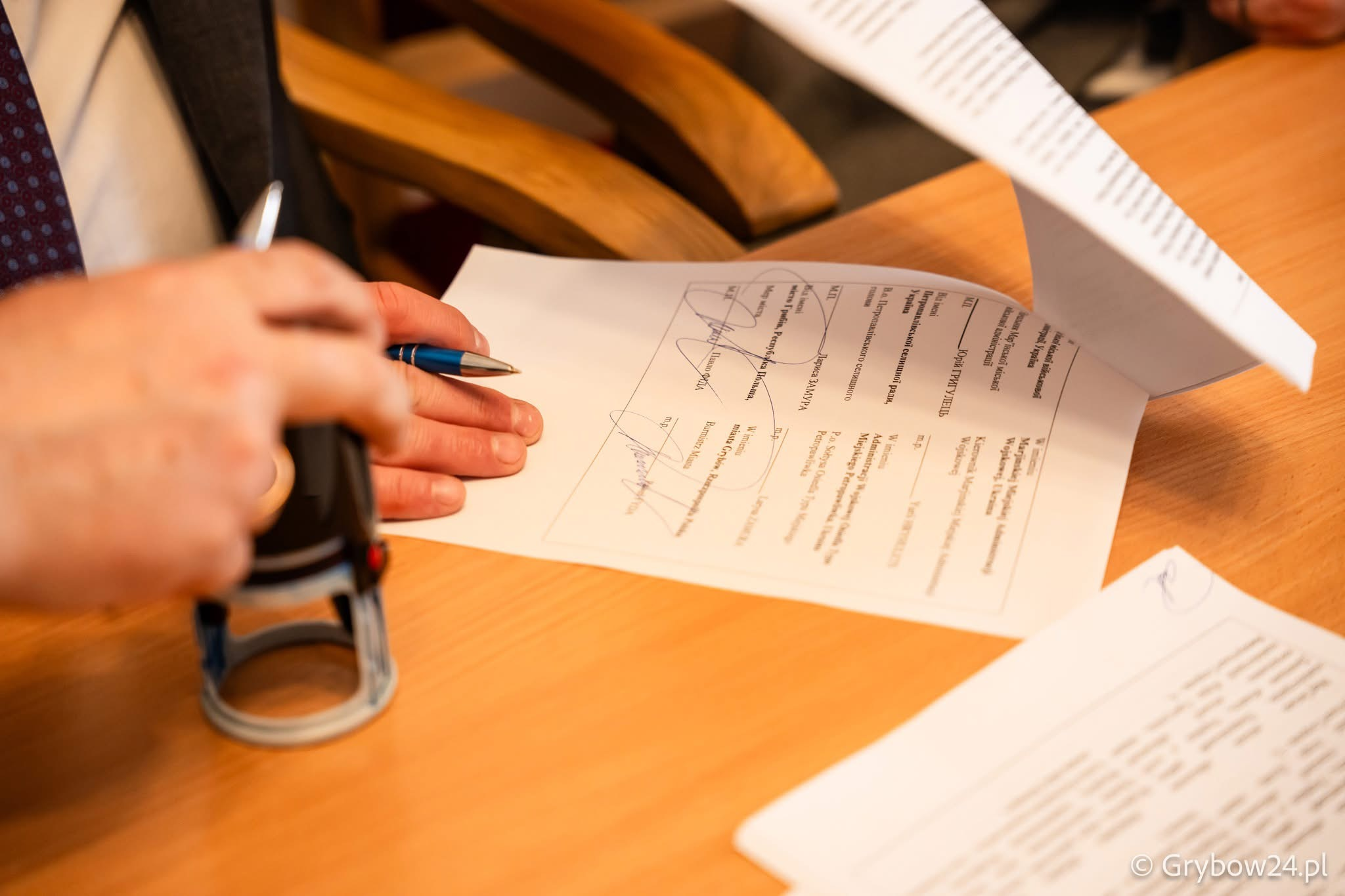 Крупний план рук за дерев'яним столом під час завірення документів. Одна рука тримає ручку, інша ставить круглу печатку на аркуш із підписами.