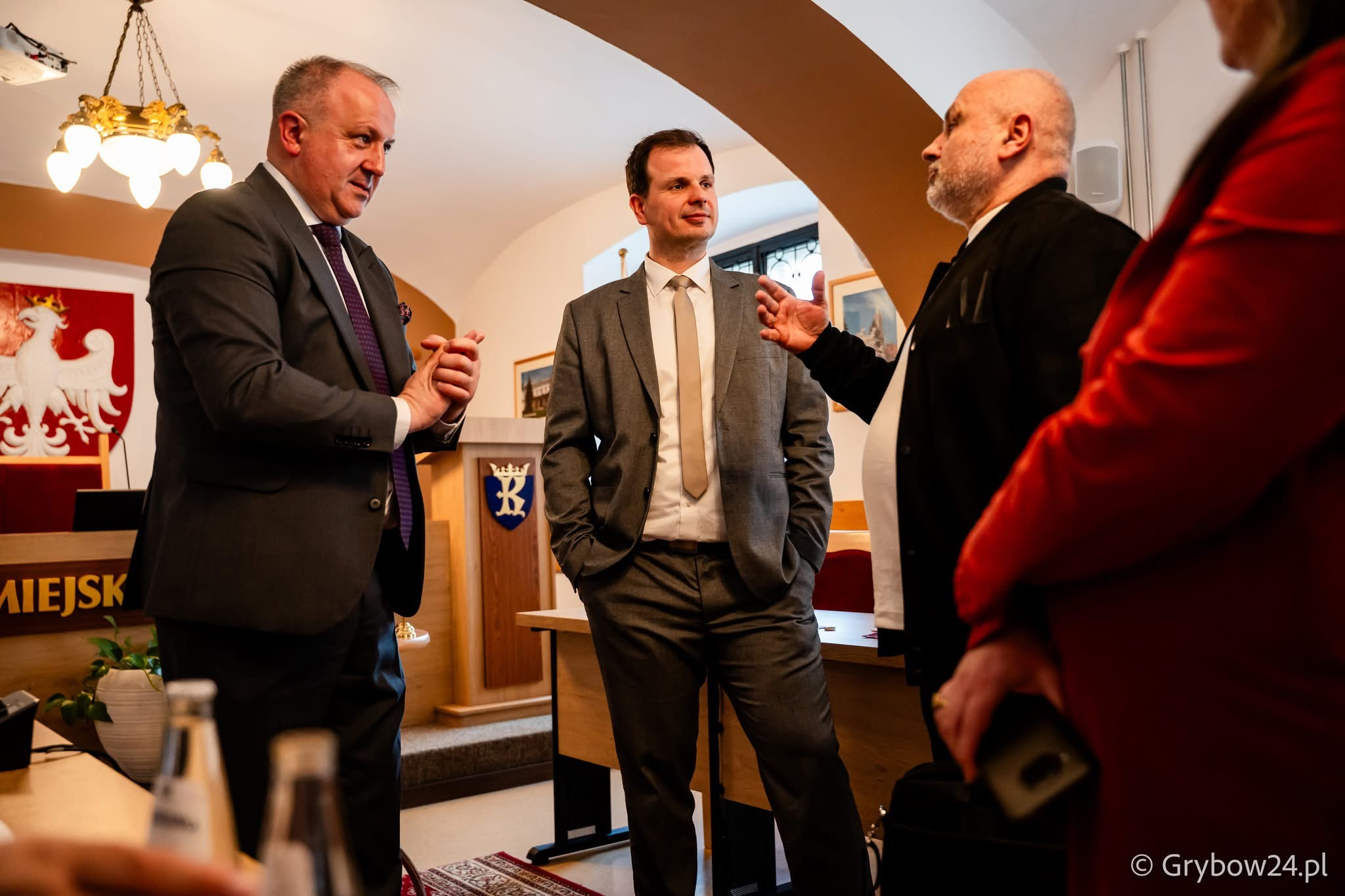 Четверо людей спілкуються в офіційному залі. Троє чоловіків у ділових костюмах стоять у центрі, один із них активно жестикулює. Праворуч на передньому плані видно частину постаті четвертої особи в червоному одязі. На фоні розміщено герб Польщі та кафедру.