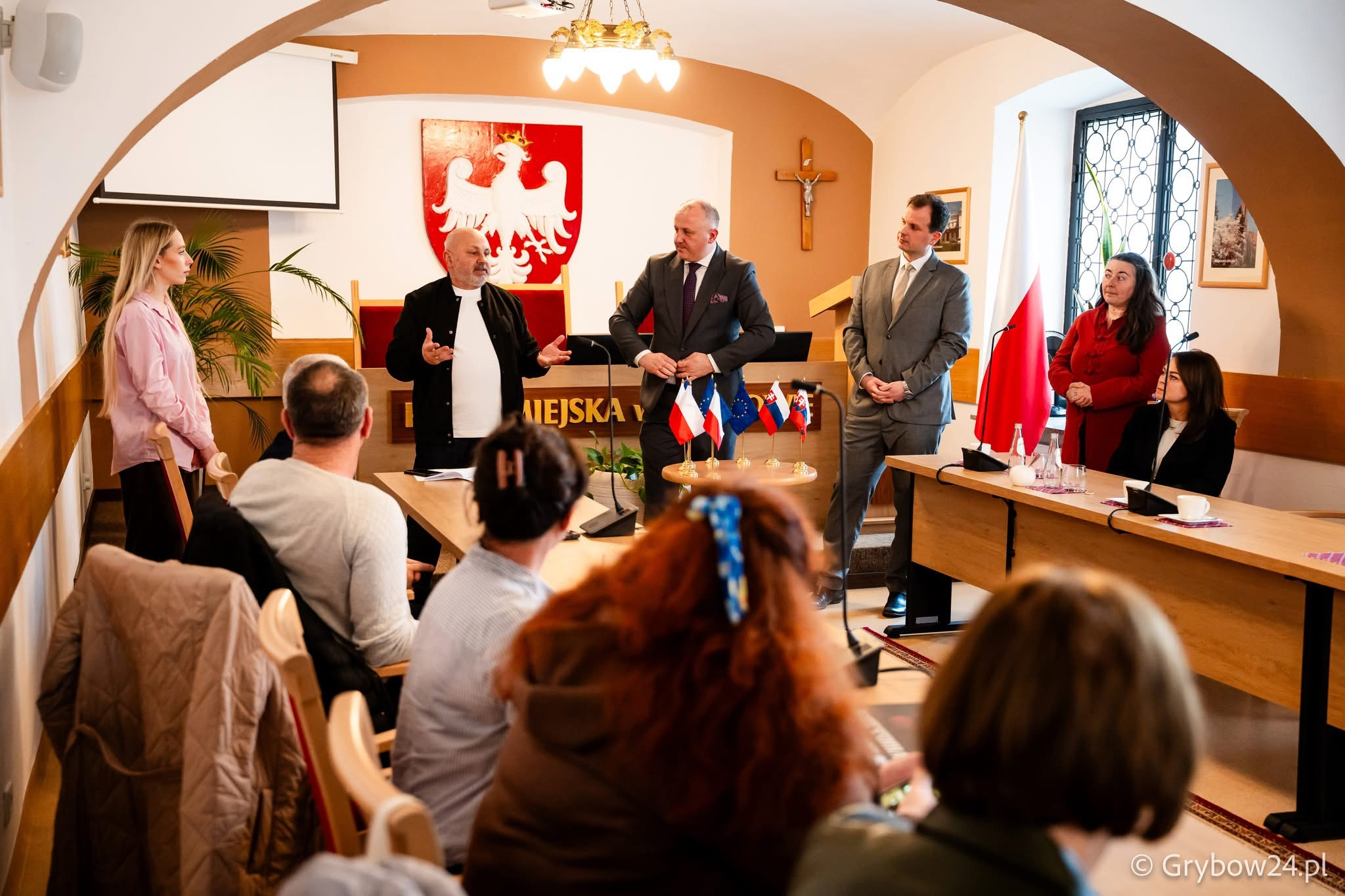 Група людей на офіційній зустрічі в залі з арочним склепінням. У центрі троє чоловіків ведуть обговорення біля столу з малими прапорами, а інші присутні сидять або стоять навколо. На стіні позаду розміщено великий герб Польщі та розп'яття.