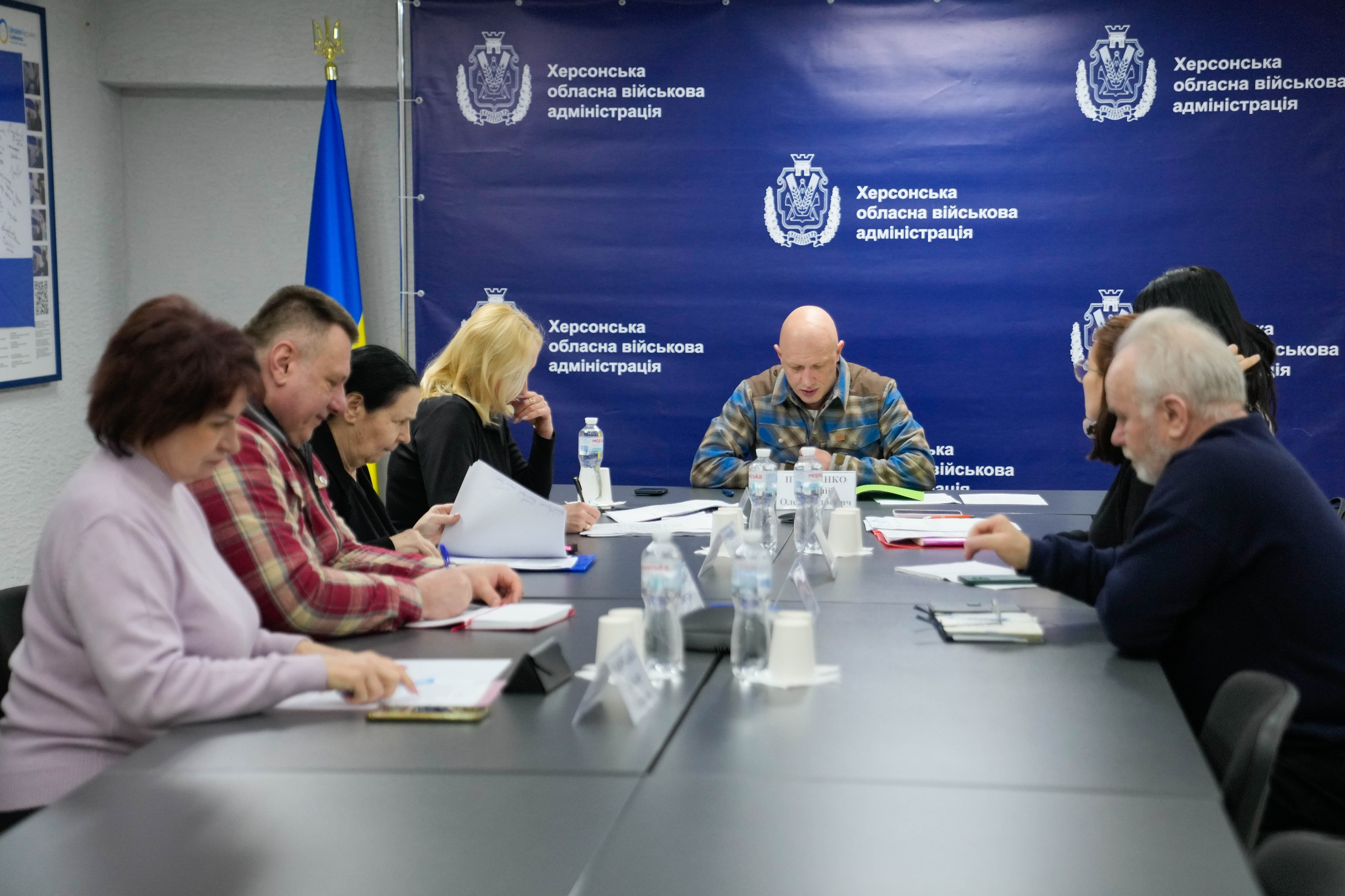 Група людей сидить за довгим столом під час наради в офіційному приміщенні. На передньому та середньому планах учасники зустрічі розглядають документи. На синьому фоні позаду них розміщено логотипи Херсонської обласної військової адміністрації, а ліворуч стоїть прапор України.