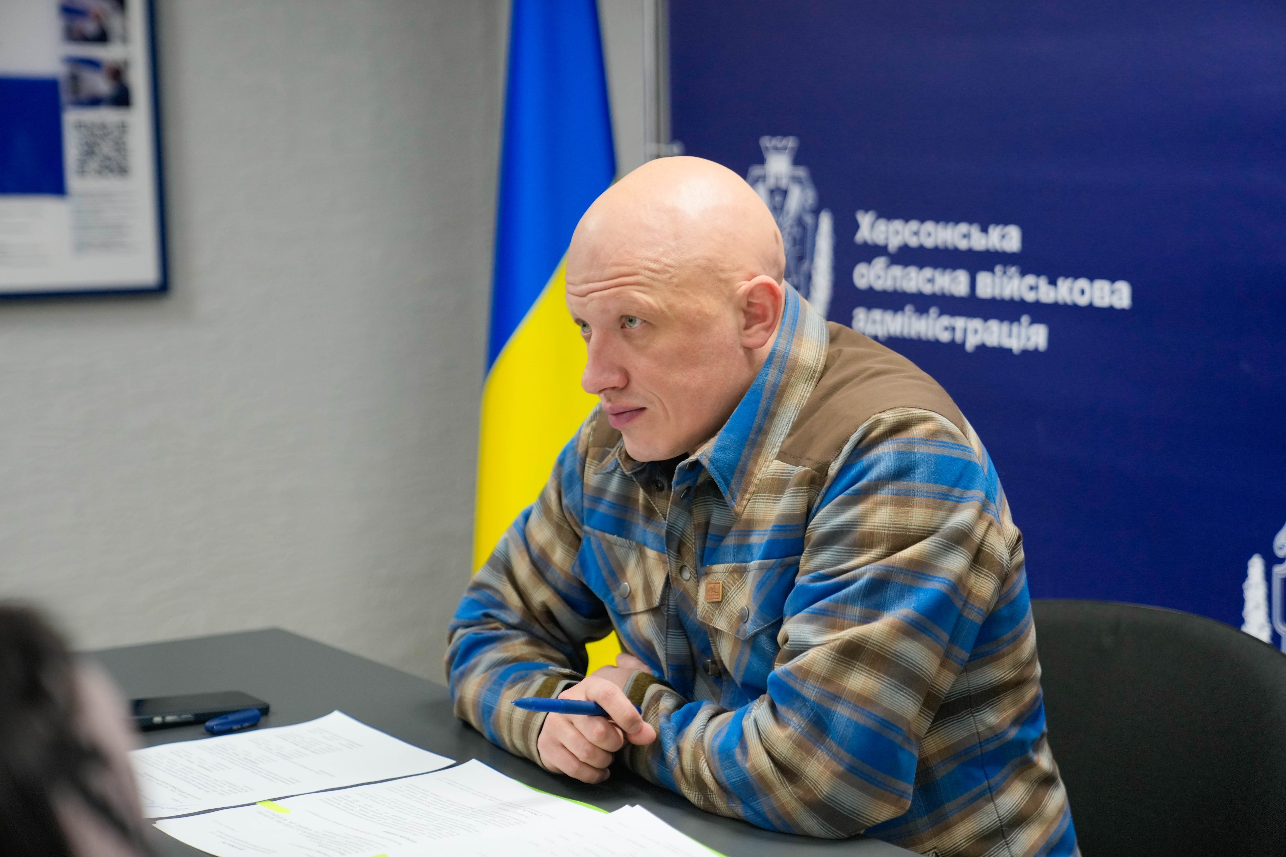 Чоловік у картатій сорочці сидить за робочим столом із ручкою та документами. На фоні за ним — прапор України та синя панель із написом «Херсонська обласна військова адміністрація».