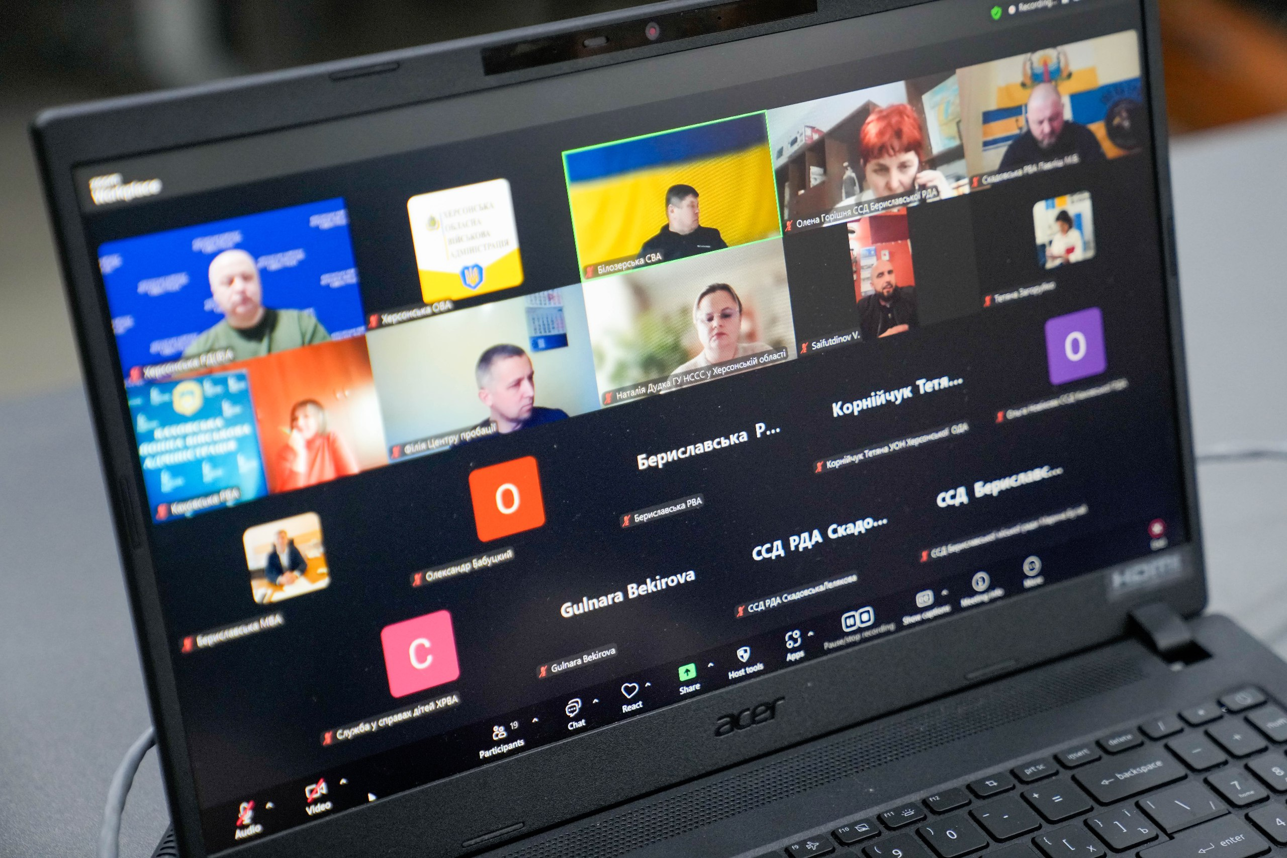 На екрані ноутбука Acer відкрито вікно відеоконференції Zoom із багатьма учасниками. Частина людей присутня з увімкненими камерами на фоні української символіки, інші відображаються як іконки з іменами та прізвищами.