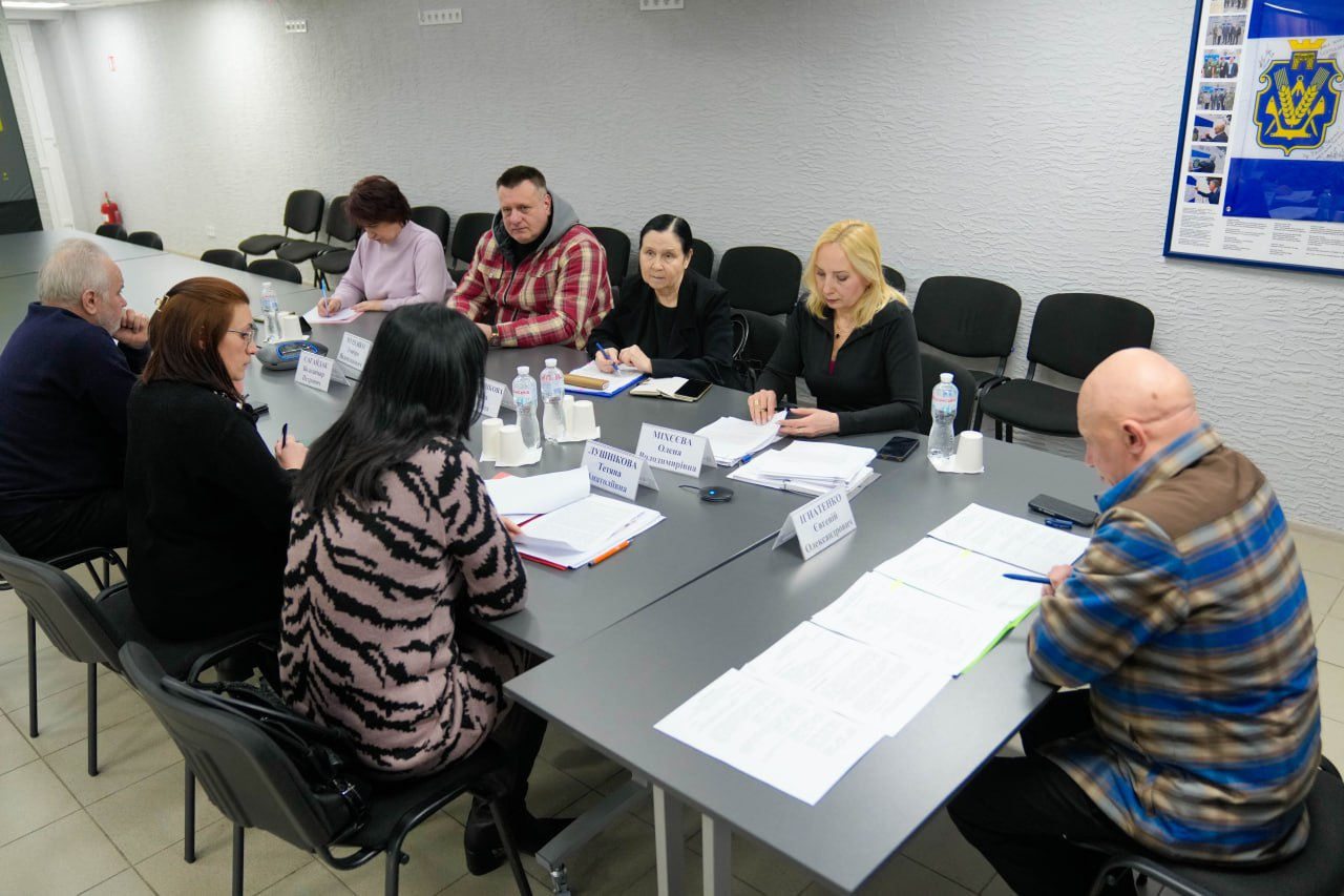 Група людей сидить за великим сірим столом під час наради або офіційної зустрічі. Учасники розглядають документи, на столі стоять пляшки з водою та таблички з іменами. На світлій стіні позаду розміщено плакат із гербом Херсонської області та іншою інформацією.