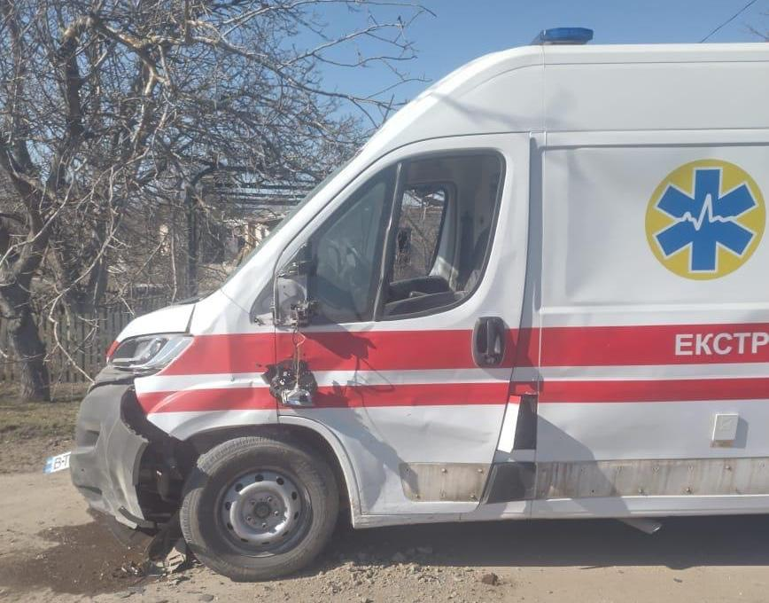 Пошкоджений автомобіль швидкої допомоги білого кольору з червоною смугою та емблемою «зірка життя». У машини розбита передня частина, відірваний бампер, пошкоджене бокове скло та вирване дзеркало. Авто стоїть на узбіччі на фоні дерев та залишків будівель.