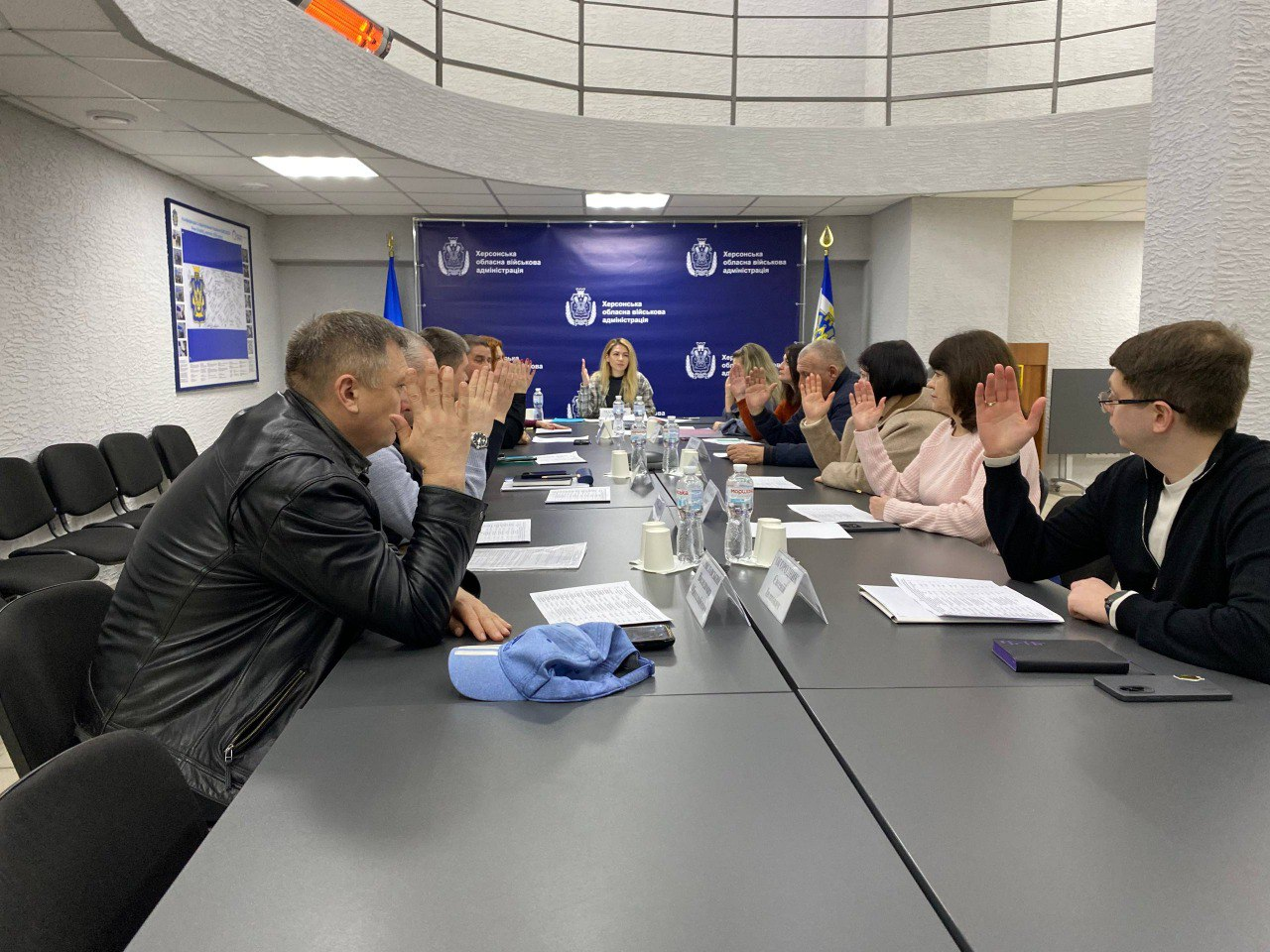 Робоча нарада в Херсонській обласній військовій адміністрації: група людей сидить за довгим столом у конференц-залі, дехто тримає підняту руку під час голосування. На фоні — синій банер із гербом та прапори України.