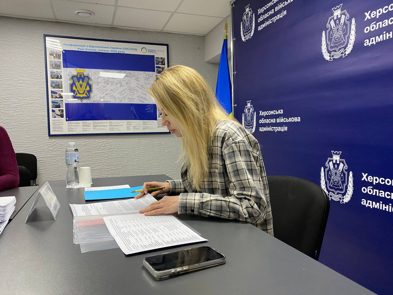Жінка в картатій сорочці працює з документами за столом у приміщенні Херсонської обласної військової адміністрації. На фоні — синій брендований банер та плакат із символікою України.