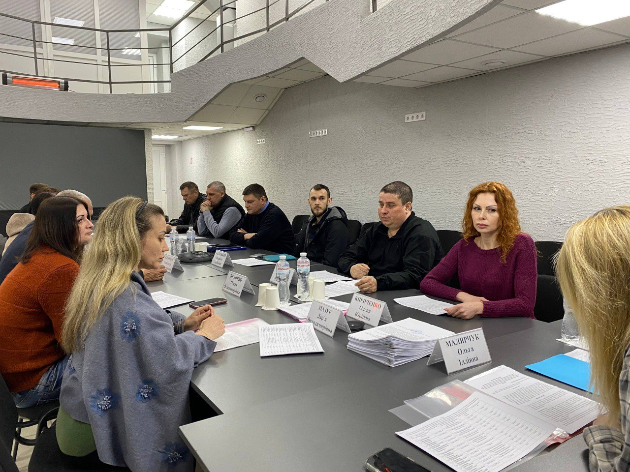 Група людей сидить за довгим столом під час офіційної зустрічі чи засідання в сучасному офісному приміщенні. Перед учасниками лежать папери, документи та іменні таблички, на столі стоять пляшки з водою.