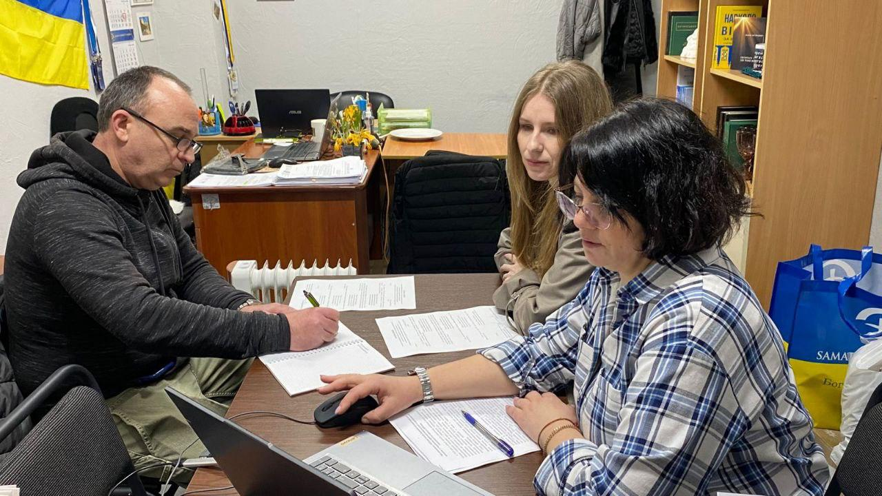 Чоловік та дві жінки сидять за столом в офісі, зосереджено працюючи з документами та ноутбуком. На задньому плані видно державний прапор України та відкритий стелаж із книгами.