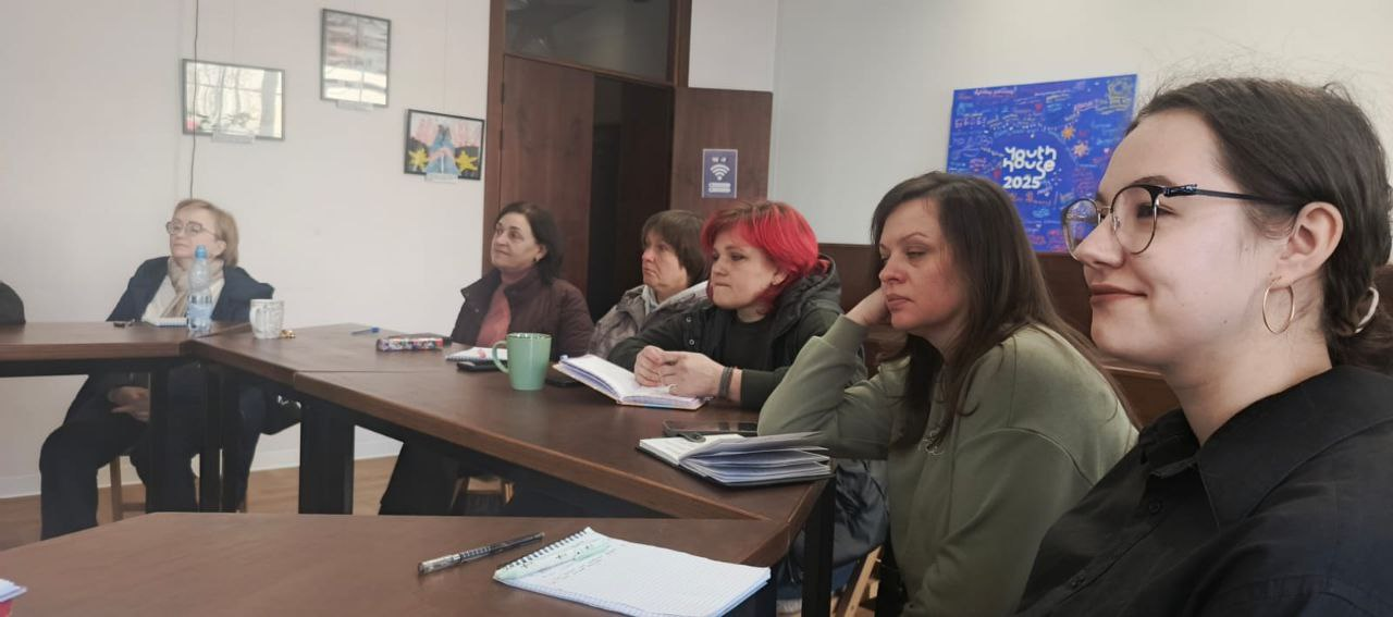 Група жінок сидить за столами під час наради чи тренінгу в офісному приміщенні. Учасниці зосереджено слухають, на столах лежать блокноти для записів, ручки та чашки. На стіні на задньому плані видно синій плакат із написом «Youth House 2025».