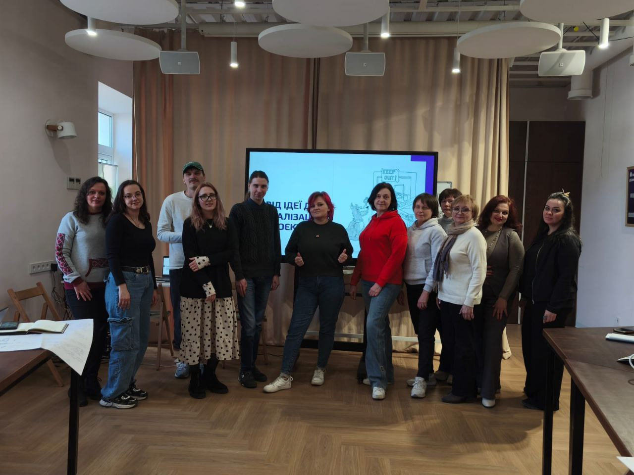 Групове фото учасників заходу в сучасному офісному просторі. Чоловіки та жінки позують перед великим цифровим екраном, на якому відображено слайд презентації. Приміщення має світлі стіни та дерев'яну підлогу, а на задньому плані видно світлі штори.