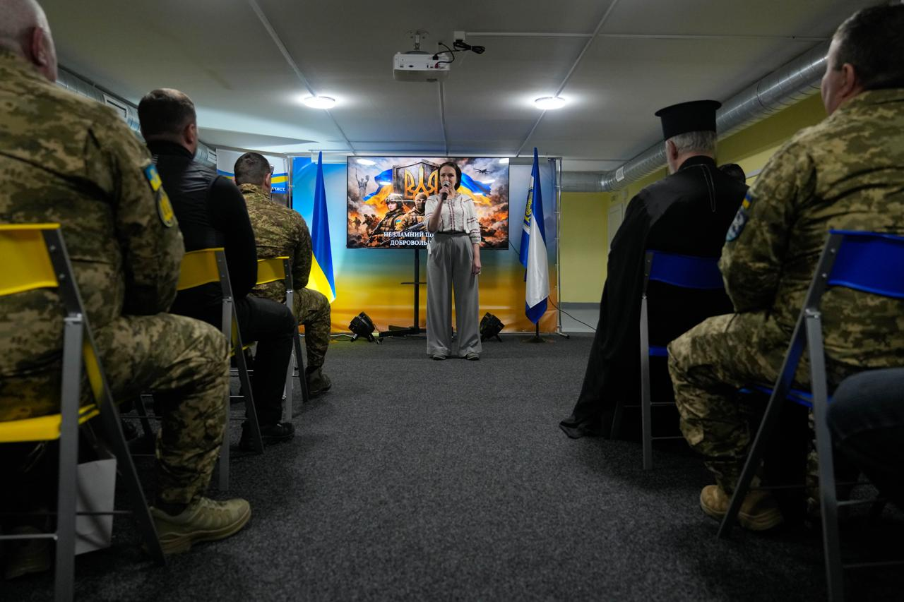 Жінка виступає з промовою перед аудиторією військовослужбовців та священнослужителя у залі. На фоні розташований великий екран із патріотичним зображенням до Дня українського добровольця та державні прапори. Захід проходить у лаконічно облаштованому приміщенні з низькою стелею.