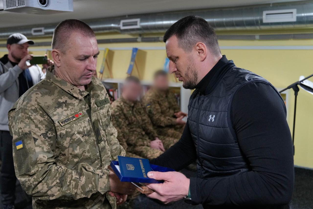 Урочиста церемонія нагородження у приміщенні. Чоловік у цивільному одязі вручає військовослужбовцю у піксельному камуфляжі нагрудний знак у синьому відкритому футлярі та посвідчення. На фоні видно інших військових та державні прапори.