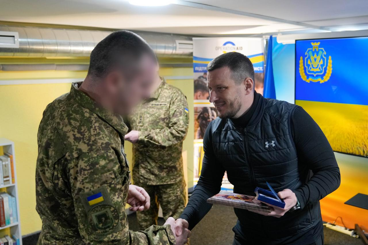 Чоловік у цивільному одязі тисне руку військовослужбовцю у піксельному камуфляжі, вручаючи йому пам'ятну табличку та нагороду у синьому футлярі. Обличчя захисника розмите з міркувань безпеки. На фоні — прапор України та екран із гербом Херсонської області.