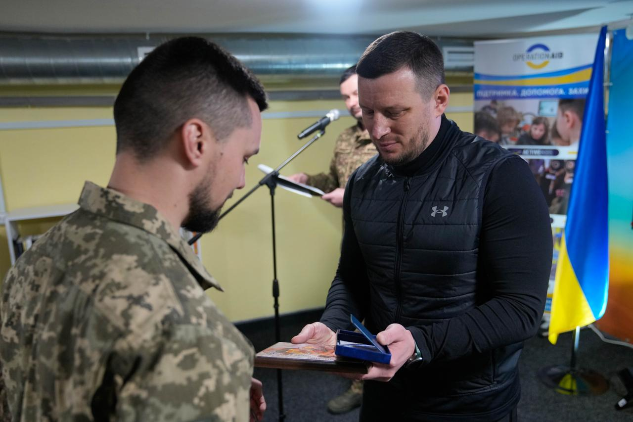 Момент нагородження захисника в офіційній обстановці. Чоловік у цивільному одязі передає військовослужбовцю у піксельному камуфляжі пам'ятну табличку та відкритий футляр із нагрудним знаком. На фоні видно Державний Прапор України та мікрофон.