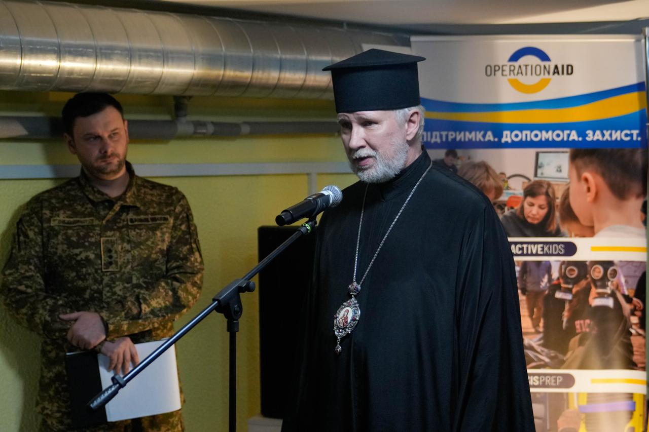Священнослужитель виступає з промовою перед мікрофоном у приміщенні. Поруч стоїть чоловік у військовому камуфляжі з папкою в руках. На фоні розташований інформаційний стенд гуманітарної організації Operation Aid із гаслом «Підтримка. Допомога. Захист».