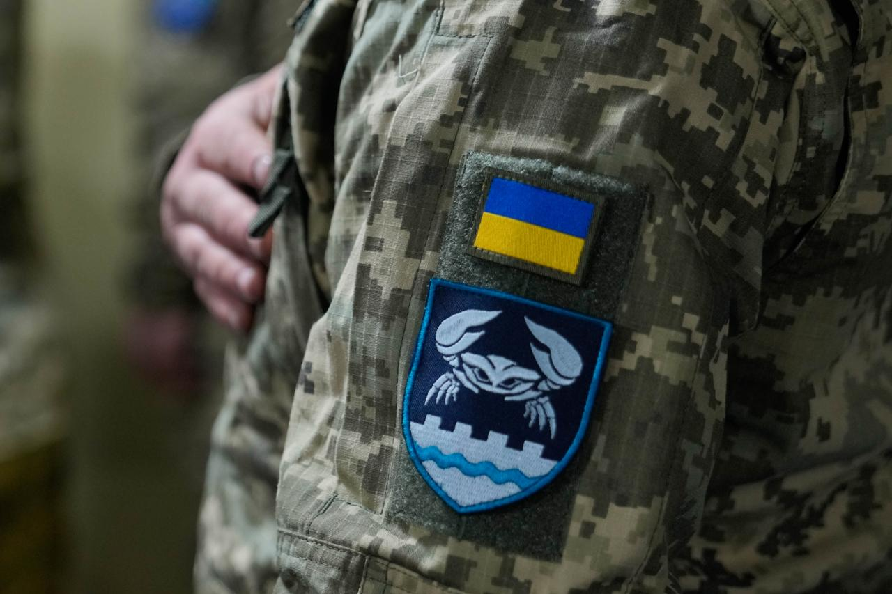 Нарукавний шеврон військовослужбовця Збройних Сил України з гербом Херсона та зображенням краба на синьому тлі. Вище розміщено невеликий нашивний прапор України. Військовий одягнений у камуфляжну форму піксельного зразка.