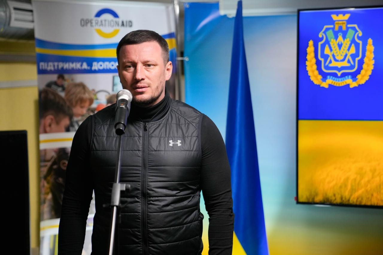 Чоловік у чорному спортивному жилеті виступає перед мікрофоном в офіційному приміщенні. Позаду нього — прапор України та великий екран із гербом Херсонської області. Ліворуч видно інформаційний стенд гуманітарної організації Operation Aid.