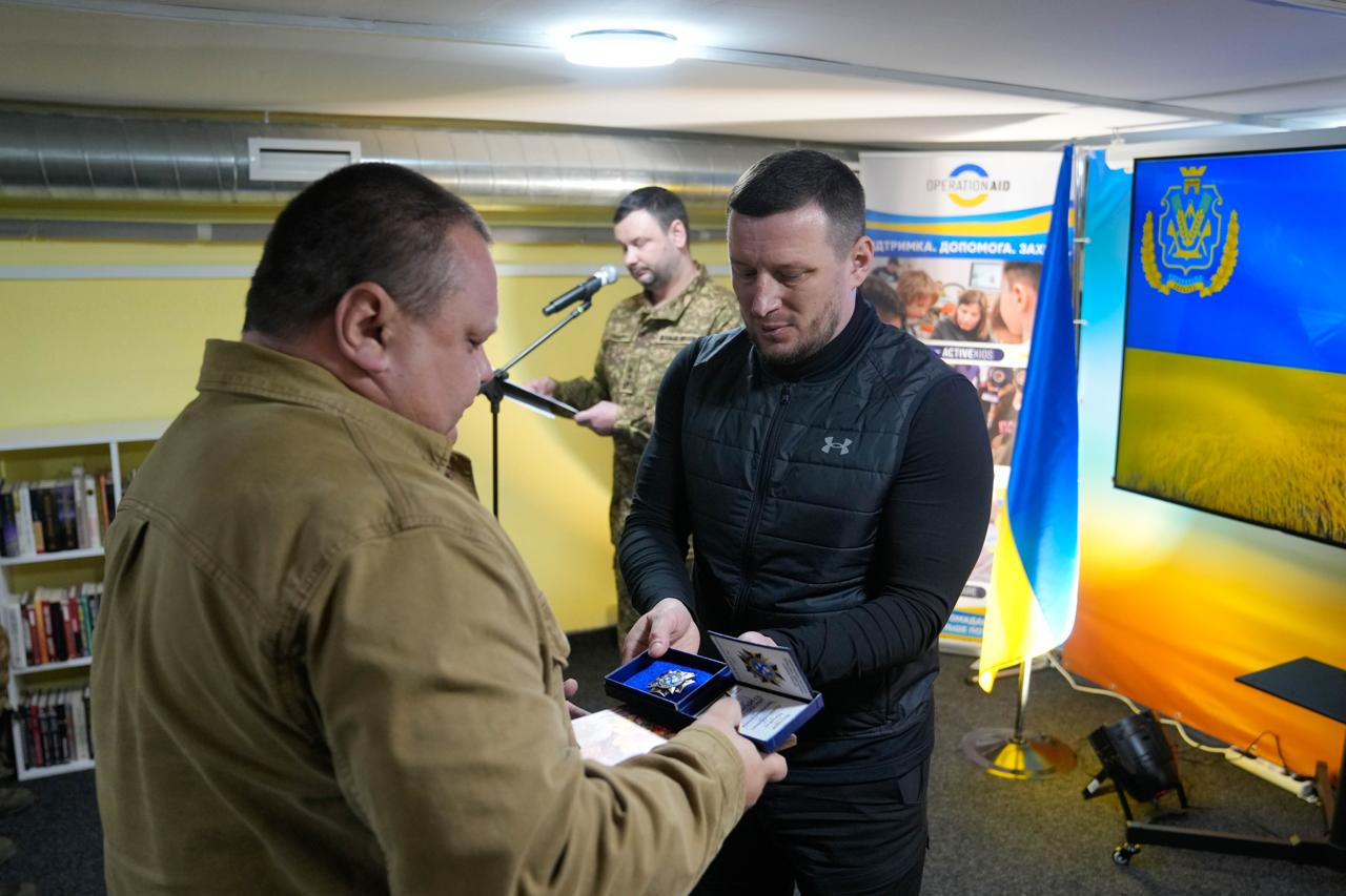 Процес нагородження в офіційній залі. Чоловік у чорному жилеті вручає нагрудну відзнаку у синьому футлярі та посвідчення іншому чоловікові. На фоні видно військового за трибуною з мікрофоном, прапор України та екран із гербом Херсонської області.