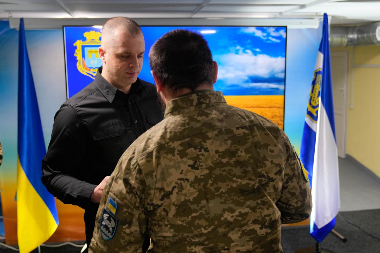 Церемонія нагородження в офіційній залі. Чоловік у чорній сорочці вручає відзнаку військовослужбовцю у піксельному камуфляжі, який стоїть спиною до камери. На фоні — Державний Прапор України, синьо-білий прапор та великий екран із гербом Херсонської області на тлі пшеничного поля.