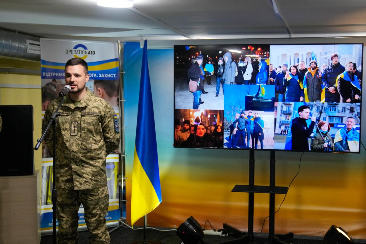 Військовослужбовець виступає з промовою біля мікрофона в офіційному залі. Поруч — прапор України та великий екран із колажем фотографій патріотичних акцій. На фоні видно банер гуманітарної організації «Operation Aid».