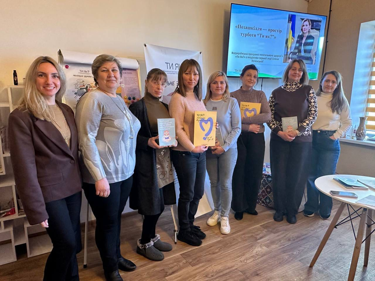 Група жінок під час презентації Всеукраїнської програми ментального здоров'я «Ти як?». Учасниці тримають інформаційні брошури з логотипом програми. На фоні — телевізійний екран із презентацією на тему «Позашкілля — простір турботи "Ти як?"».